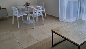 A modern living room with a round dining table, white chairs, a flat-screen TV mounted on the wall, and a large window with sheer curtains. - Le Paradis à la Plage (Linguizzetta)