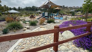 A playground with a climbing structure, a covered seating area, and a garden with purple flowers and rocks.
