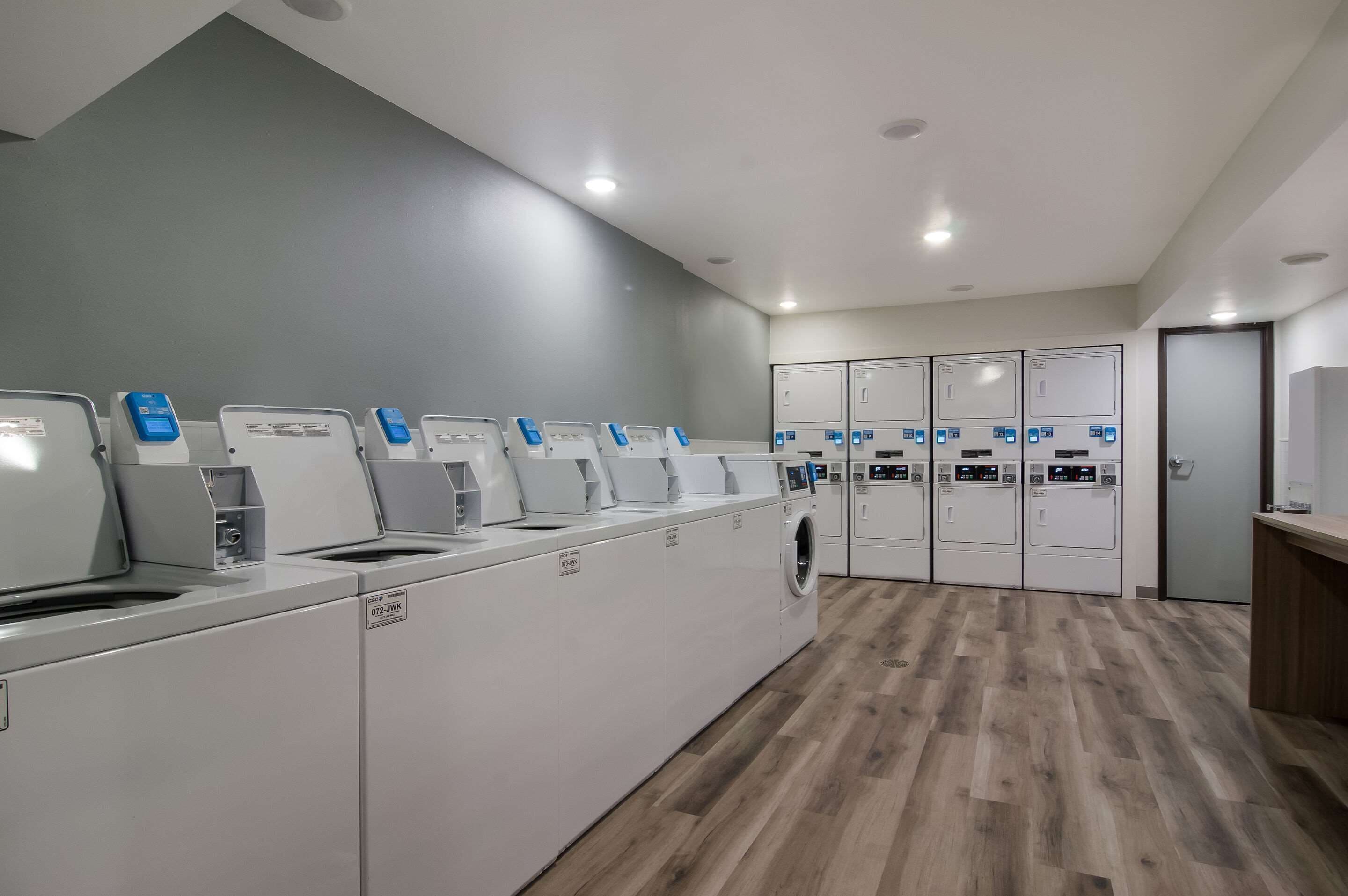 A laundry facility with multiple washing machines and dryers, a wooden floor, and a grey wall.