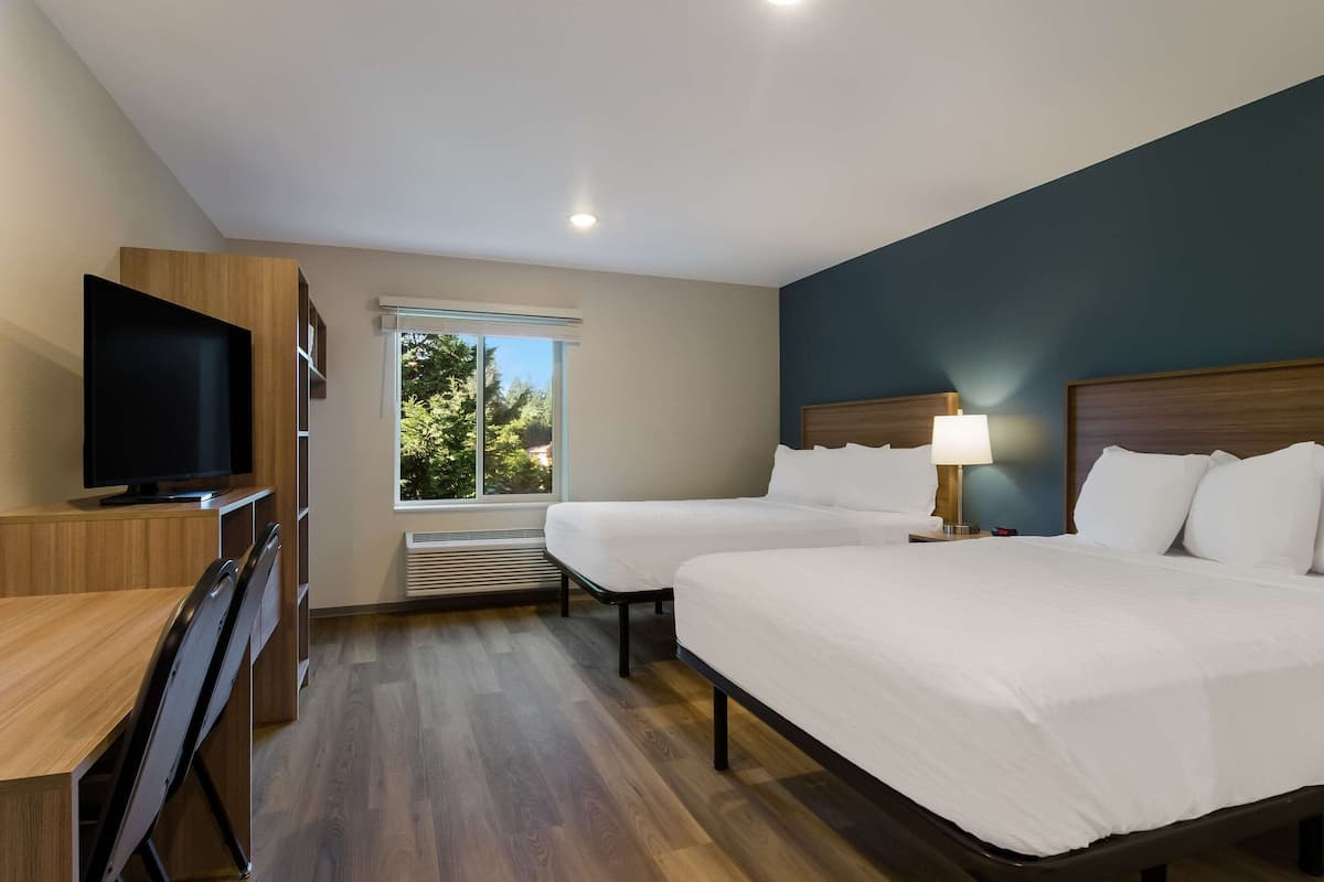 A hotel room with two beds, a TV, a desk, and a window with a view of trees.