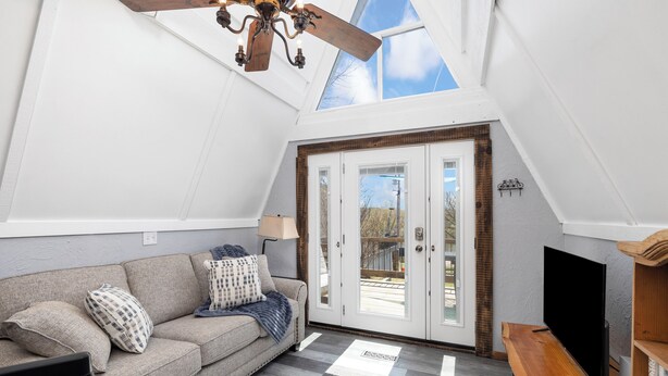 Un salon chaleureux avec un plafond haut, un ventilateur de plafond en bois, un canapé avec des coussins, un téléviseur posé sur un meuble en bois et une grande porte vitrée donnant sur l’extérieur.