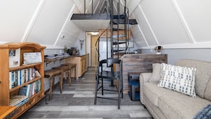 Un appartement de style « loft » avec une bibliothèque en bois, un escalier en colimaçon en métal noir, un canapé agrémenté de coussins à motifs et un coin cuisine.