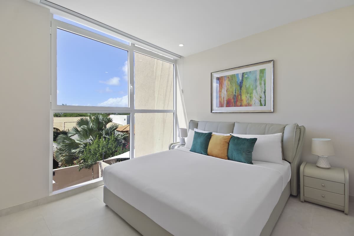 A modern bedroom with a large bed, a nightstand, a framed abstract painting, and a view of palm trees through a large window.