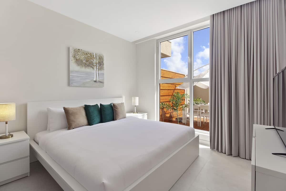 A modern hotel room with a large bed, a nightstand with a lamp, a painting on the wall, and a view of the outdoors through a window.