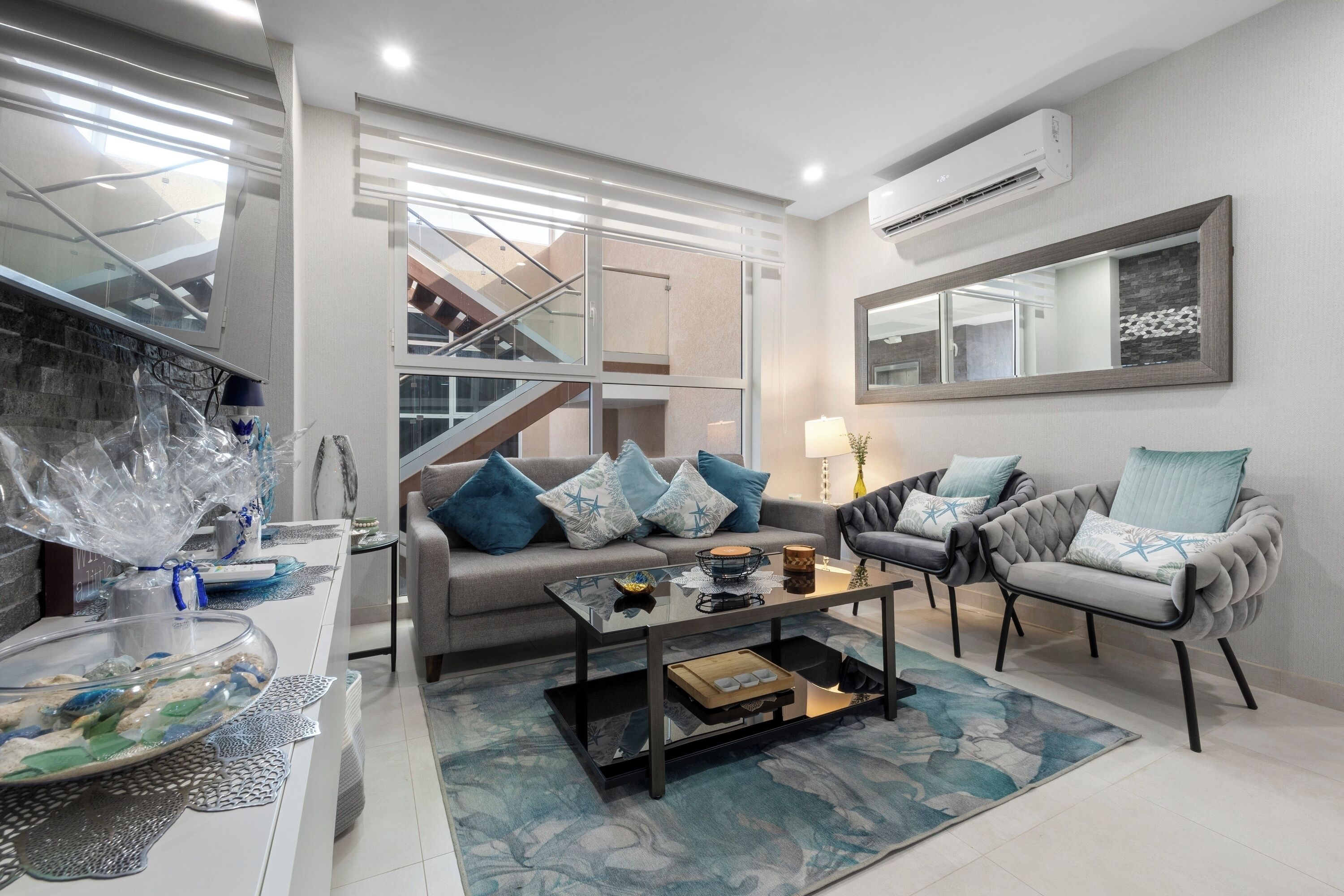 A modern living room with a sofa, coffee table, and decorative items. A staircase is visible in the background, leading to an upper floor.