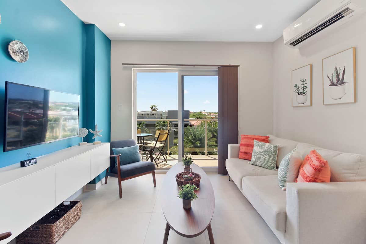 A modern living room with a flat-screen TV, a white sofa, a coffee table with plants, and a balcony with a view.