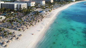 Un resort en la playa con varios edificios hoteleros, una playa de arena bordeada de palmeras y sombrillas, y aguas turquesas cristalinas.
