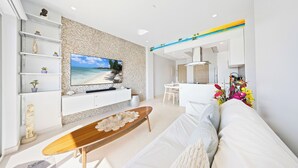 Una sala de estar moderna con un televisor de pantalla plana, sofás blancos, una mesa de centro de madera y una cocina al fondo.