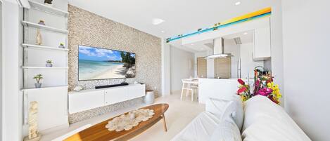 A modern living room with a flat-screen TV, white sofas, a wooden coffee table, and a kitchen area in the background.