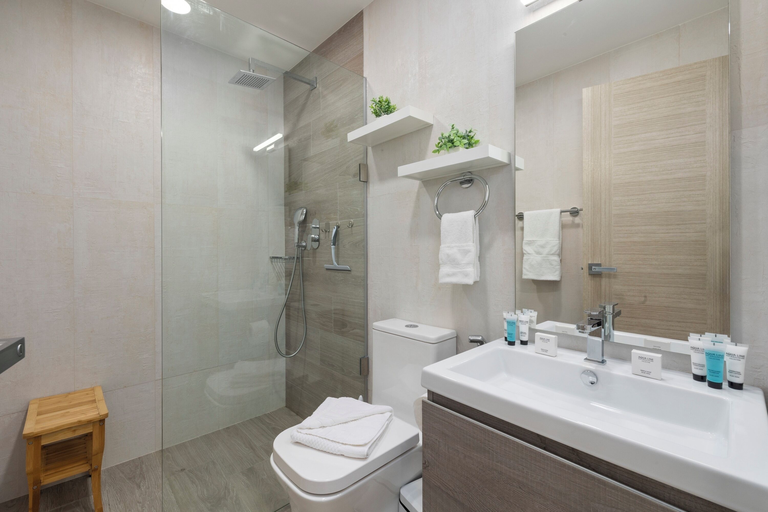 Un baño moderno con ducha de vidrio, inodoro, lavamanos con artículos de aseo y un taburete de madera.