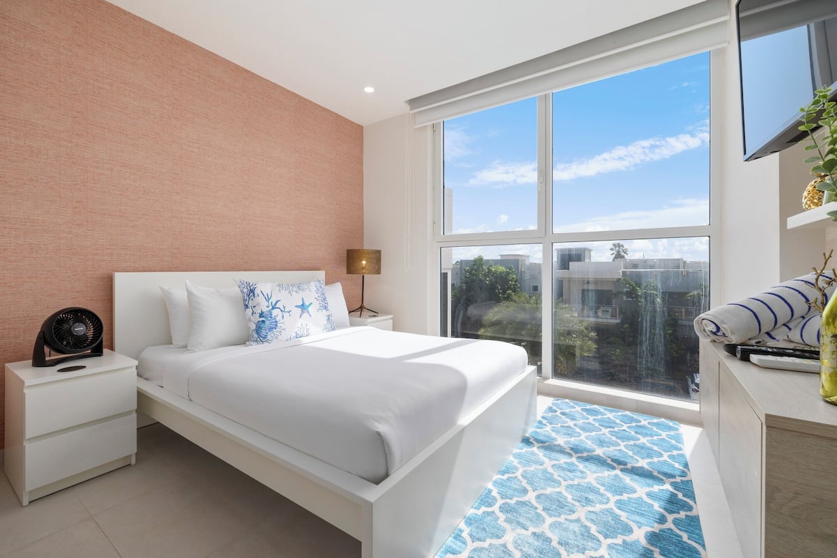 A modern bedroom with a large window, a bed with white bedding, a bedside table with a lamp, and a view of the city.