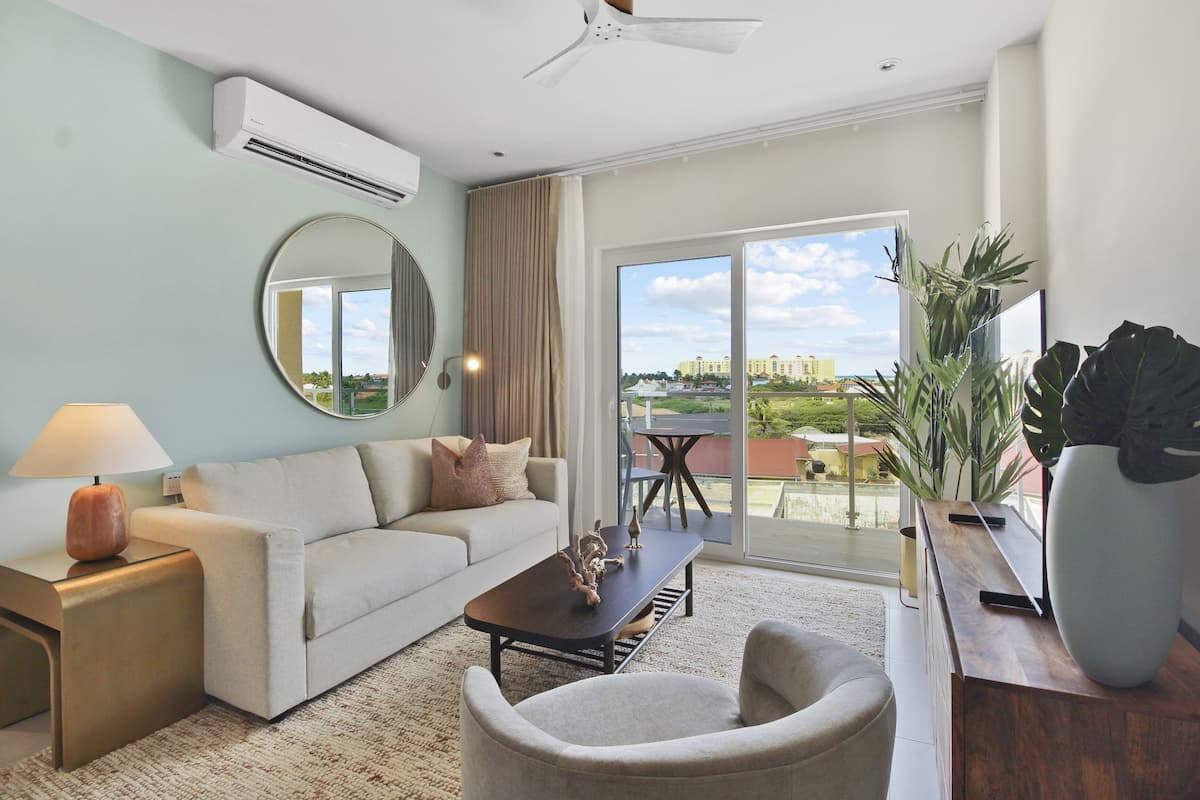 A modern living room with a sofa, coffee table, and chairs, a large mirror, and a view of a cityscape through a sliding glass door.