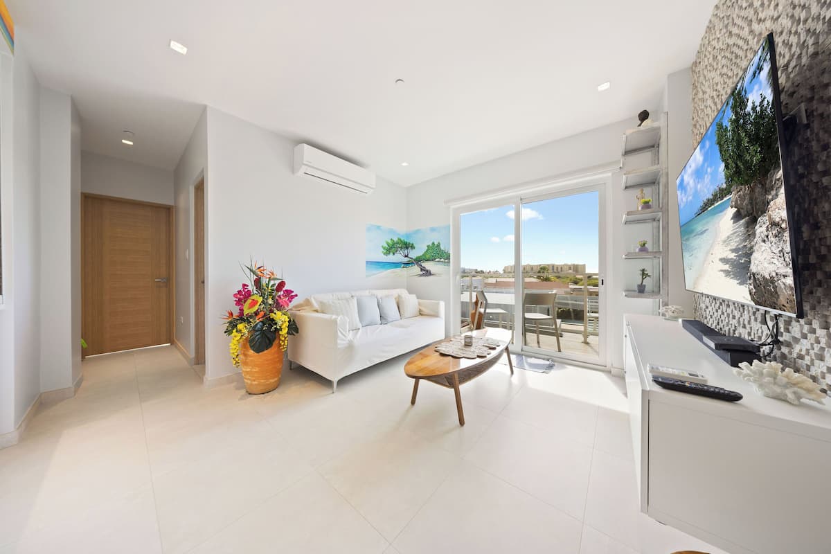 A modern living room with a white sofa, a wooden coffee table, and a large TV on a white cabinet. There is a potted plant with colorful flowers and a painting of a beach scene on the wall.