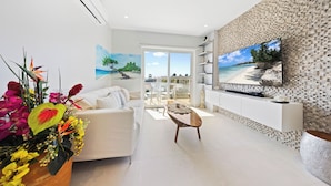 Una sala moderna con un sofá blanco, una mesa de centro y un televisor grande montado en una pared texturizada. Hay un balcón con vista al mar.