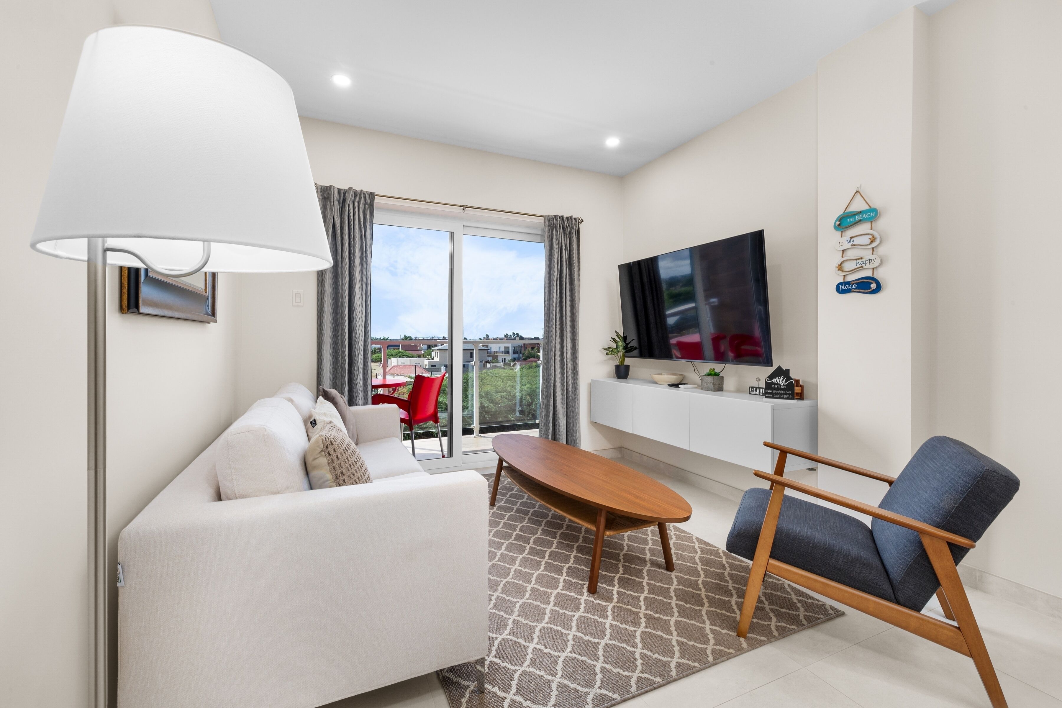 A modern living room with a sofa, a coffee table, and a TV. There is a large window with curtains, a flat-screen TV mounted on the wall, and a small table with decorative items.