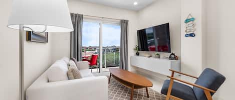 A modern living room with a sofa, a coffee table, and a TV. There is a large window with curtains, a flat-screen TV mounted on the wall, and a small table with decorative items.