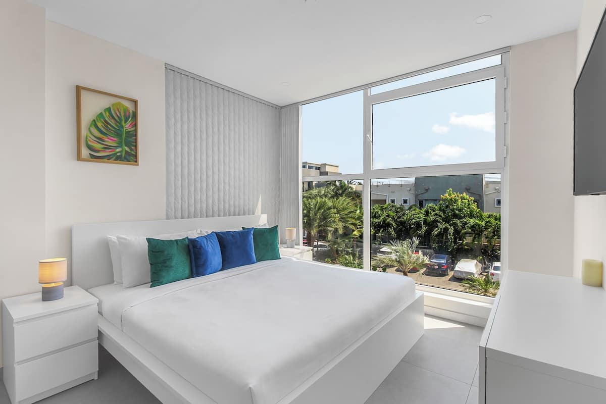 A modern hotel room with a large bed, white nightstands, a bedside lamp, and a large window offering a view of the city.
