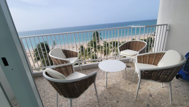En balkon med fletstole og et lille bord, med udsigt over en strand med palmer og havet.