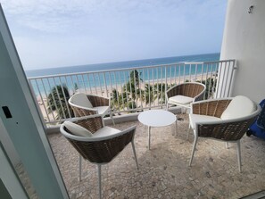 Un balcone con sedie in vimini e un tavolino, affacciato su una spiaggia con palme e l'oceano.
