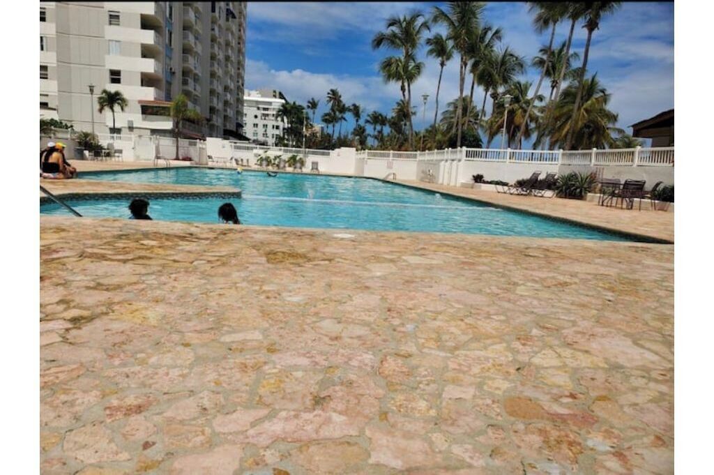 A swimming pool with stone flooring, surrounded by palm trees and a white fence.