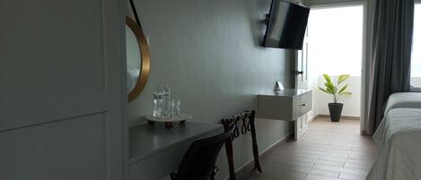 A hotel room with a bed, a desk with a chair, a TV mounted on the wall, a potted plant, and a window with curtains.