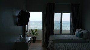 Habitación de hotel con cama, televisor montado en la pared, planta en maceta y vista al mar a través de un ventanal grande.
