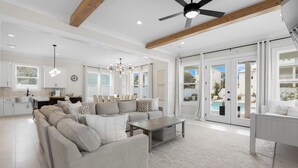 A spacious living room with a large sectional sofa, a coffee table, and a dining area with a white table and chairs. There is a kitchen area with white cabinets and a pendant light. The room has large windows and a view of a pool outside. - Seagrove Beach - Private Pool + Spa - Bikes (Seagrove)