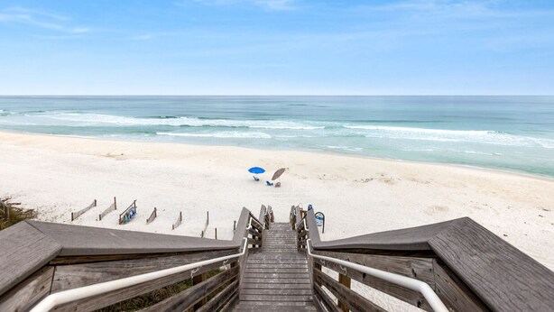 A wooden staircase leading to a beach with a clear view of the ocean, a blue umbrella, and a few beach chairs.