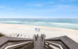 A wooden staircase leading to a beach with a clear view of the ocean, a blue umbrella, and a few beach chairs.