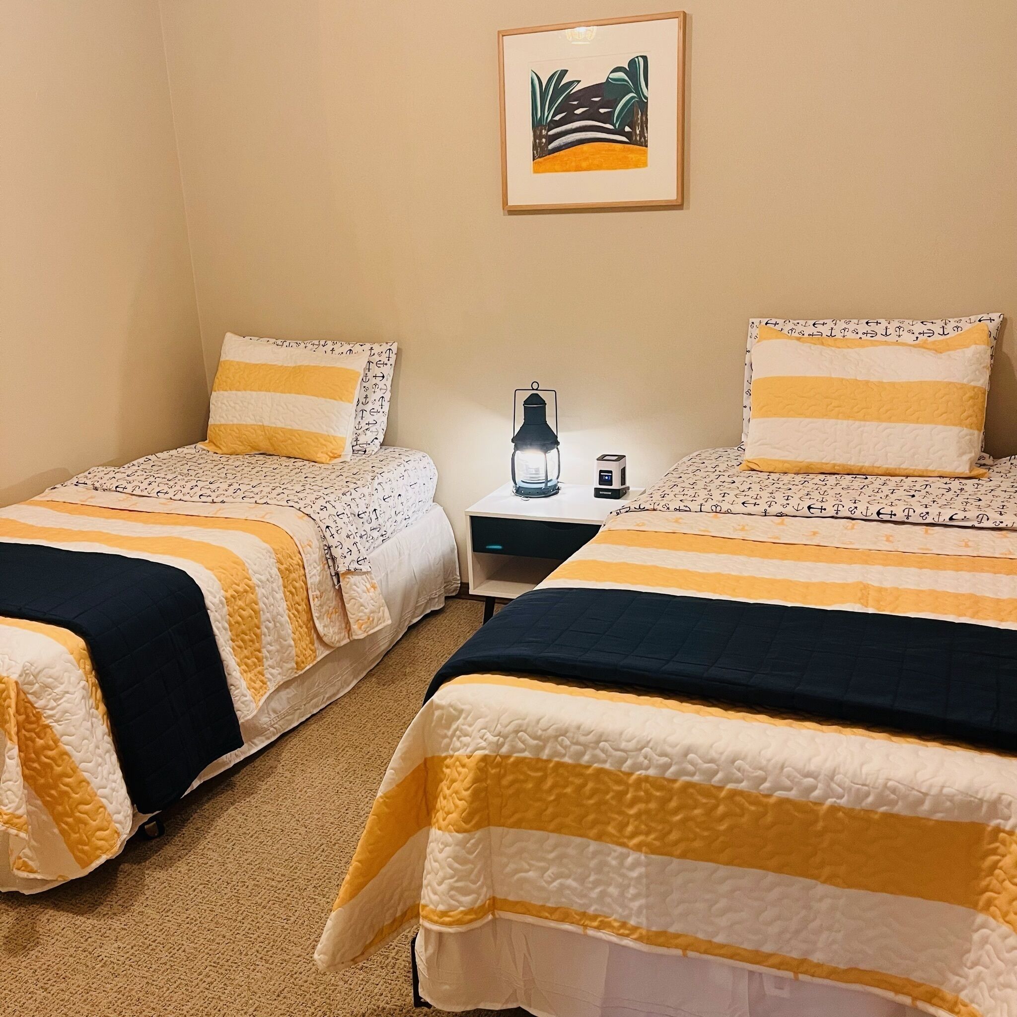 Ein Zimmer mit zwei Betten, jeweils mit gestreifter Bettwäsche, einem kleinen Tisch mit Lampe und einem gerahmten Bild an der Wand.