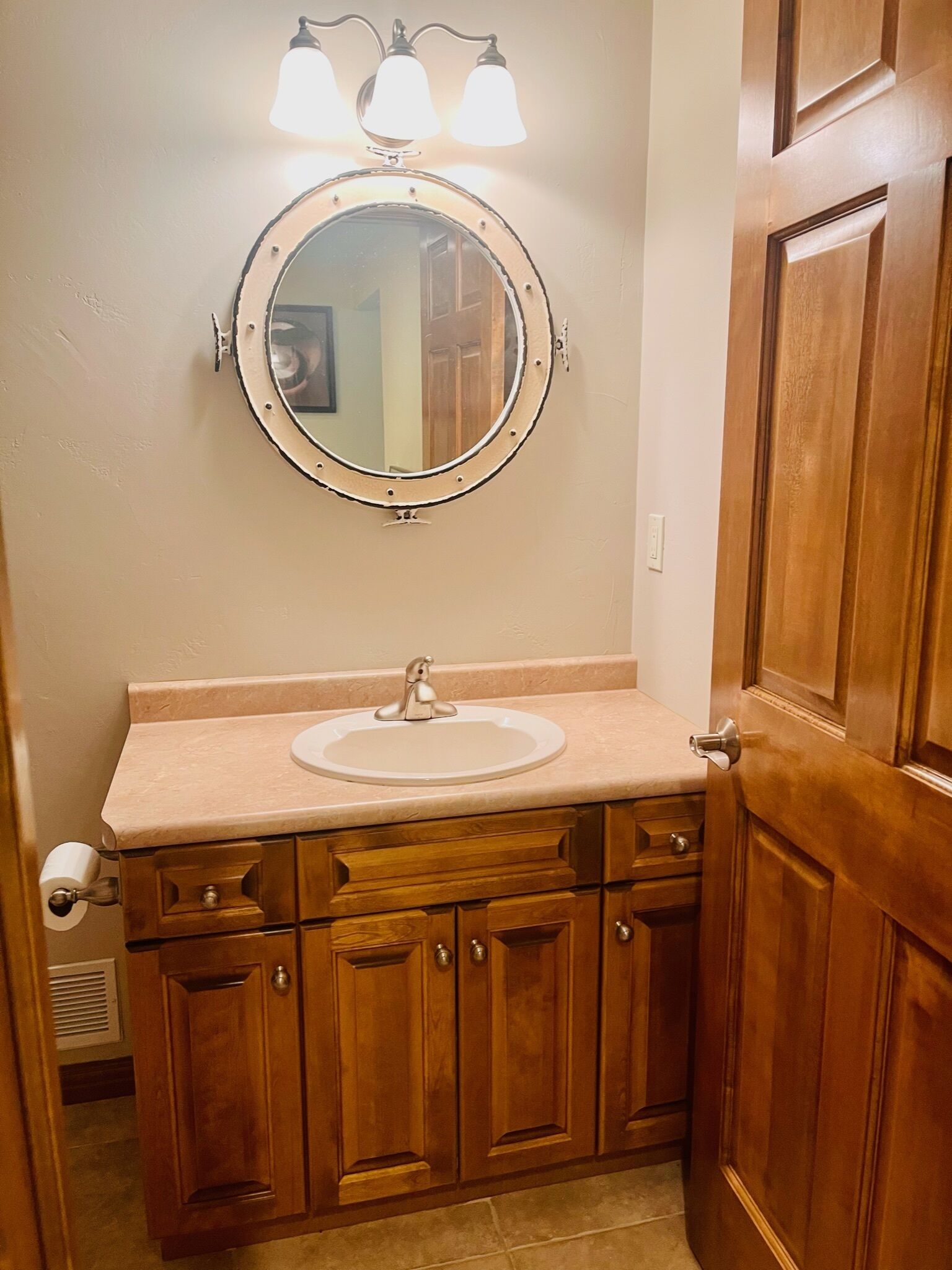 A bathroom with a wooden vanity, a round mirror with three lights, and a white sink.
