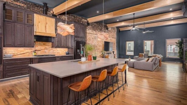 A modern kitchen with dark wood cabinets, a large island with bar stools, and a brick wall. - Luxury Loft Overlooking Downtown (Lincolnton)