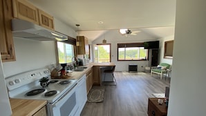 A kitchen with wooden cabinets, a stove, and a dining area with a table and chairs. - Acadia Birds Eye View Cottage (Southwest Harbor)