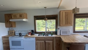 A kitchen with wooden cabinets, a stove, a sink, and a window with a view of greenery. - Acadia Birds Eye View Cottage (Southwest Harbor)