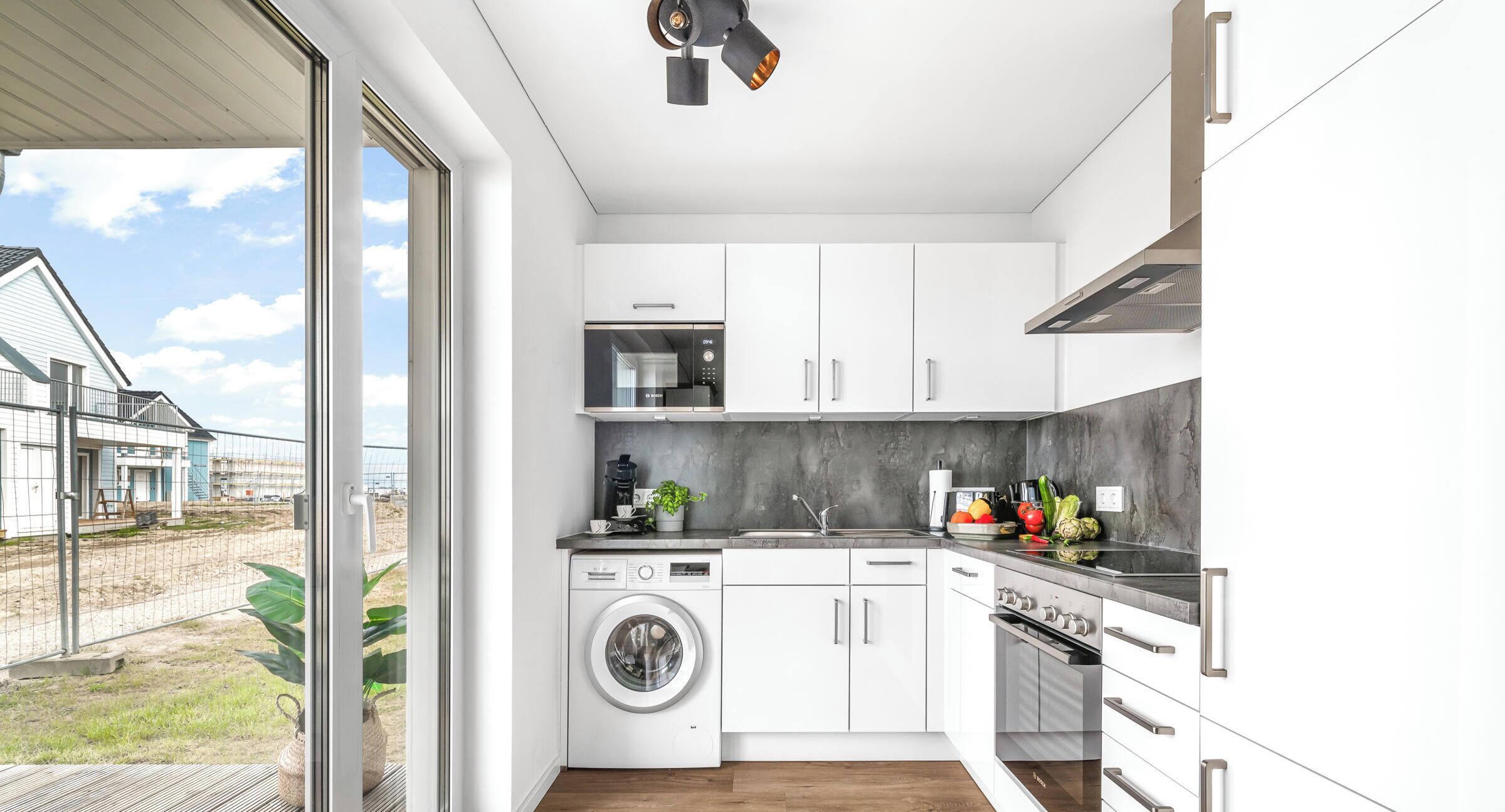 Une cuisine moderne équipée d’un lave-linge, d’un four et offrant une vue sur une maison grâce à une porte vitrée.