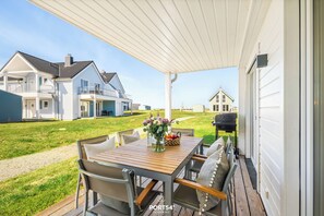 A covered patio with a wooden table and chairs, a vase with flowers, and a grill in the background.