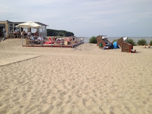 Ein Strand mit einer Holzterrasse, Stühlen, Sonnenschirmen, einer kleinen Holzkonstruktion und Sand.