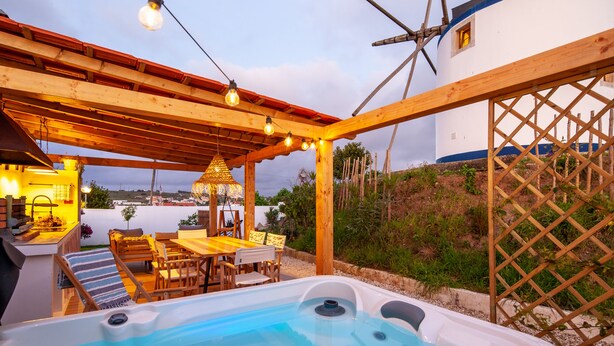 Een hottub onder een houten pergola met lichtjes, omringd door een terras met zitplaatsen en uitzicht op een wit gebouw met blauwe accenten.