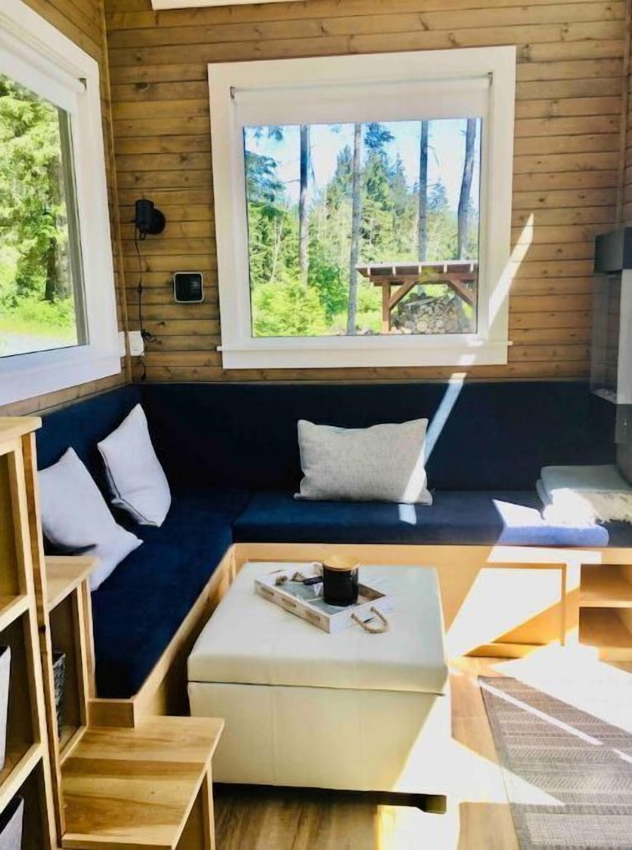 En mysig stuga med en mörkblå soffa, vita kuddar, ett träbord och ett fönster med utsikt över en skog.