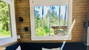 Interior acollidor d'una cabana amb un sofà blau fosc, coixins blancs, una taula de cafeteria de fusta i una finestra amb vistes a un bosc.