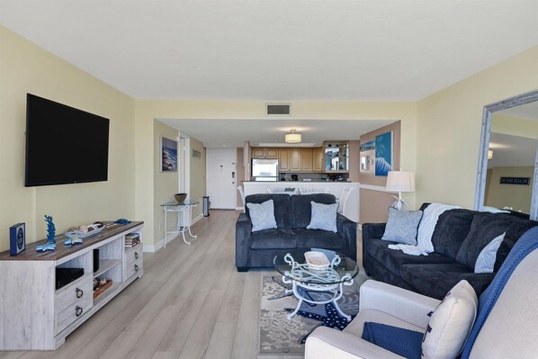 A living room with a TV, two couches, a glass coffee table, and a kitchen in the background.