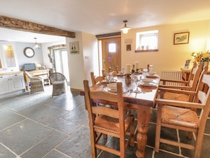 A dining area with a wooden table set for six, surrounded by wicker chairs. A kitchen with a stone floor and a wooden door is visible in the background. - Hen Dy Craig Yr Ronwy (Bala)