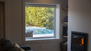 Una sala de estar con un ventanal que da a una piscina y árboles, chimenea y sofá verde.