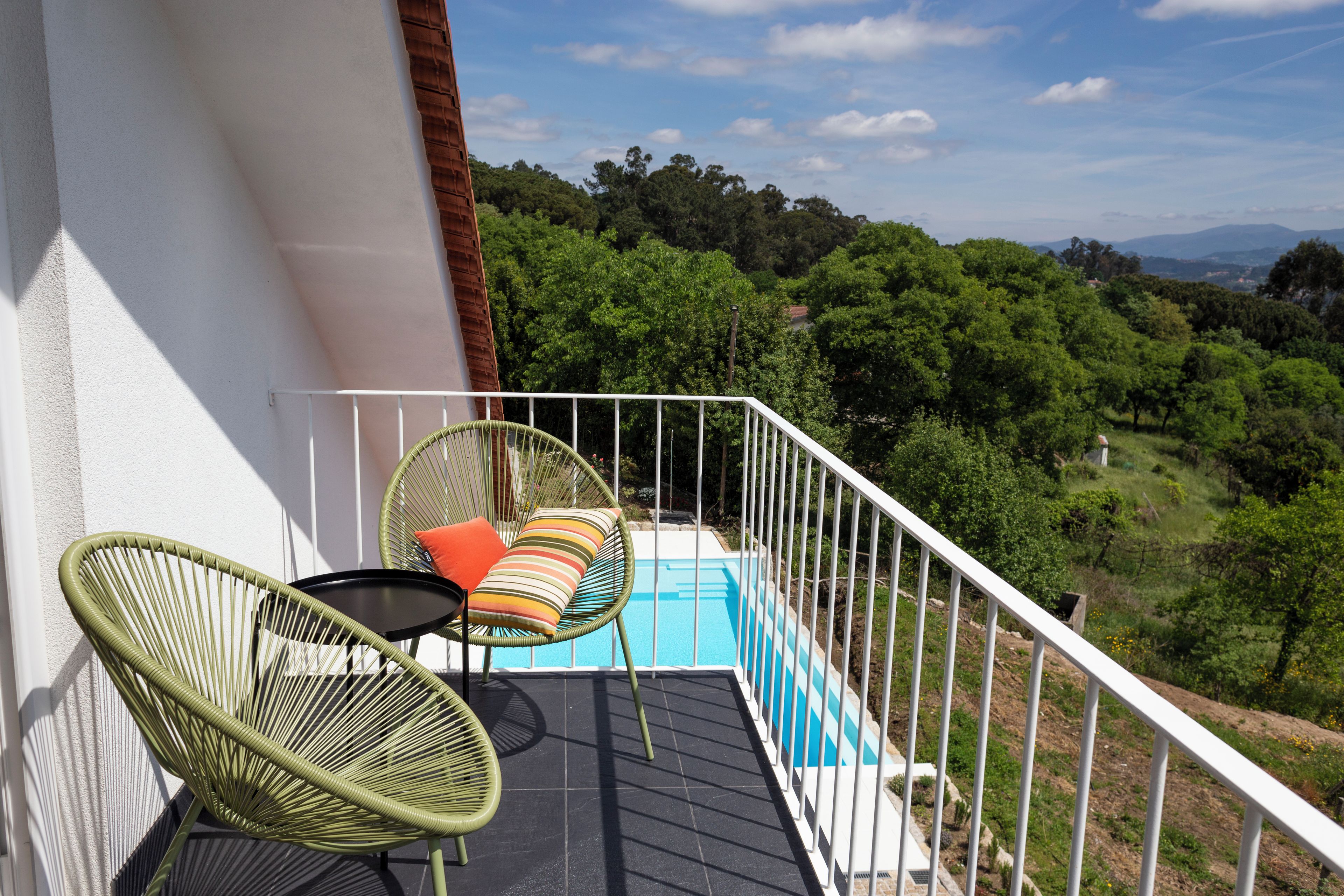 Un balcón con piscina, dos sillas de mimbre y vistas a un paisaje boscoso.