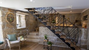 Un interior moderno con pared de piedra, una escalera de madera con baranda de metal, una silla de mimbre y una pequeña mesa con una planta.
