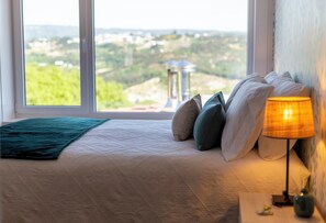 A bedroom with a large window, a bed with pillows, a bedside lamp, and a view of a distant landscape.