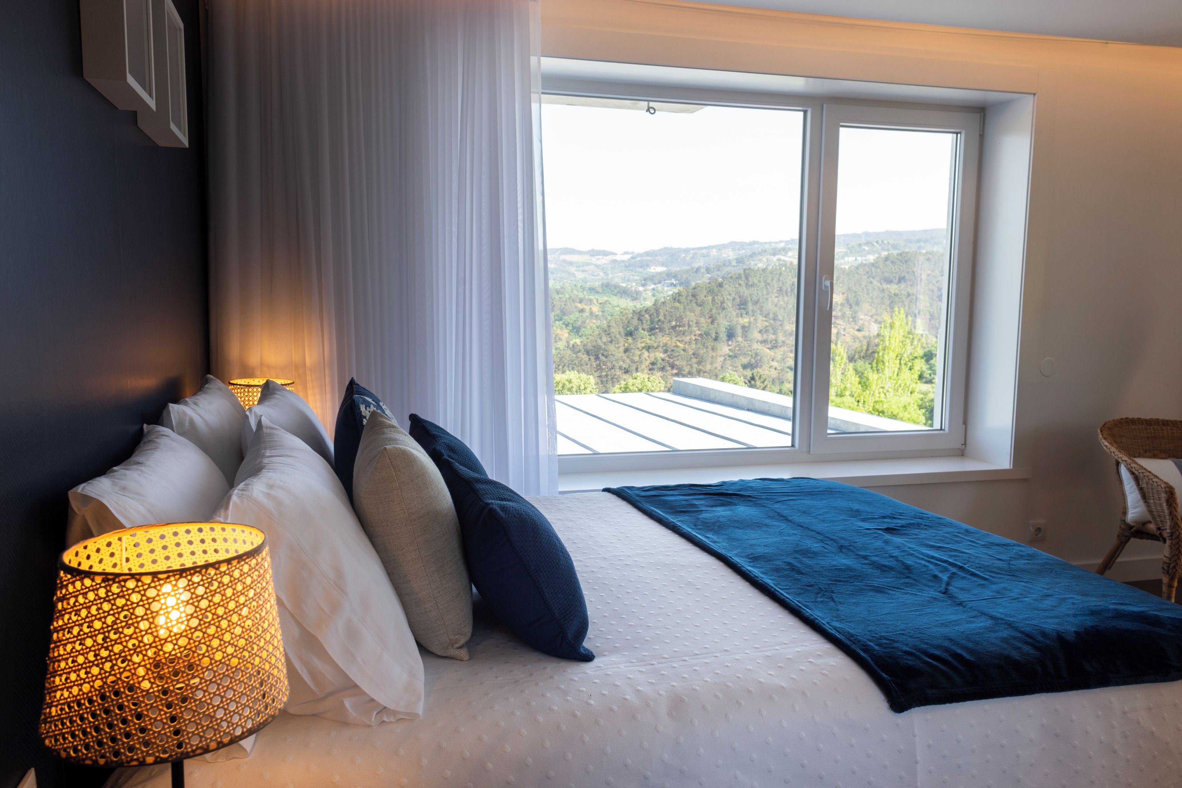 Un dormitorio con una ventana grande, una cama con almohadas, una lámpara y vistas a un paisaje boscoso.