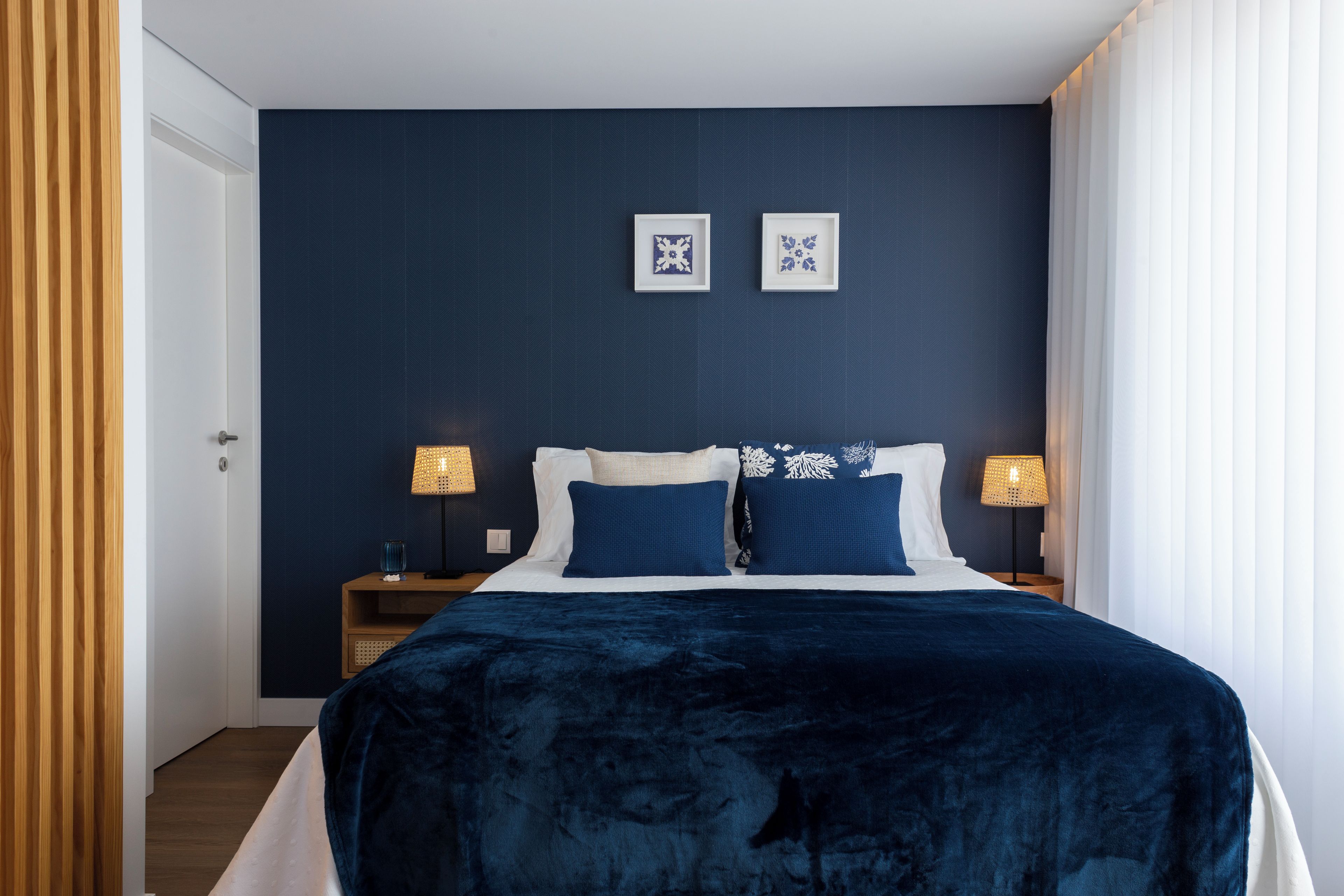 Un dormitorio con una pared azul oscuro, una cama grande con una manta azul, dos mesitas de noche con lámparas y dos cuadros enmarcados en la pared.