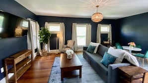 Un salon moderne avec un mur bleu foncé, une table basse en bois, un canapé agrémenté de coussins à motifs, un luminaire suspendu et un téléviseur placé sur le mur de gauche.