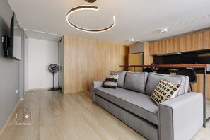 A modern living room with a grey sofa, wooden panel walls, a flat-screen TV, and a kitchen area with wooden cabinets and a black countertop.
